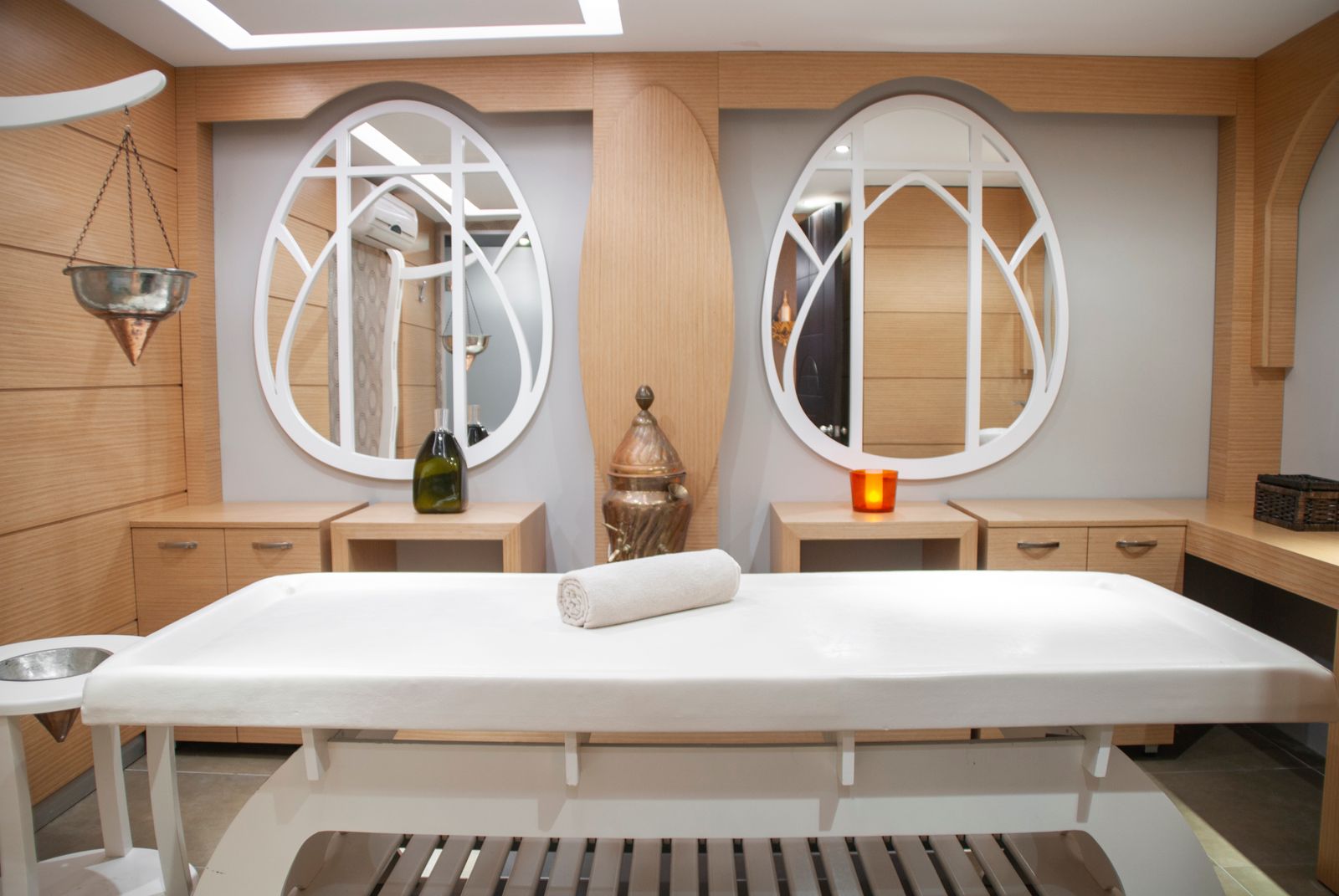 A calm spa treatment room with a white massage table and rolled towel, two large egg-shaped mirrors on a grey wall, pale wood-panelled walls, a green glass bottle and an orange candle holder.