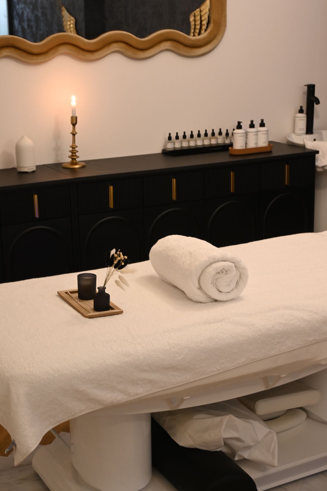 A white-towelled massage table with a rolled towel and a small wooden tray holding a dark candle and a bud vase with dried grasses, in front of a black fluted sideboard with brass handles carrying a lit candlestick and rows of small bottles, under a wavy gold-framed mirror.