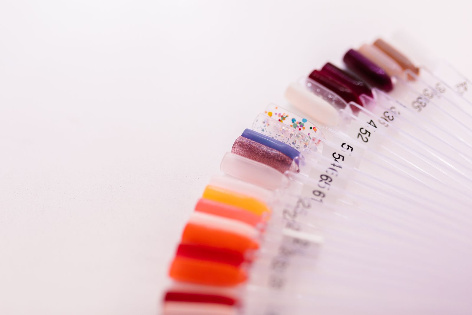 A fan of clear nail-colour sample sticks on a white surface in reds, oranges, pinks, mauve, glitter and berry tones, with small shade numbers printed on the sticks, in soft focus.