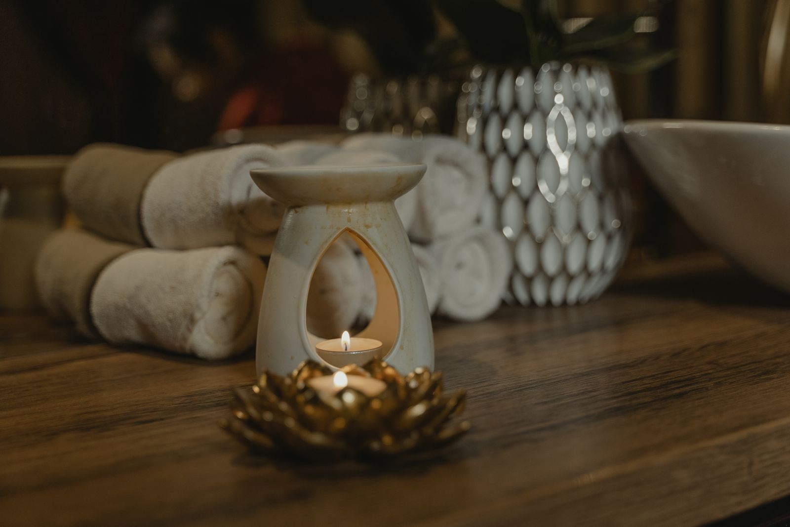 A warm close-up of a cream ceramic oil burner with a lit tealight and a gold lotus candle holder on a wooden counter, with rolled beige towels softly out of focus behind.